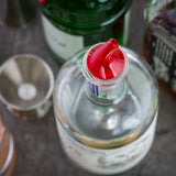 A close-up of a clear liquor bottle with a BPA free, 1 oz. pourer on top, surrounded by other bottles and a metal jigger on a gray surface. The focus is on the bottle’s neck and spout, with Proper Pour III accessories softly blurred in the background.