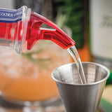 A close-up photo shows clear liquid being poured from a BPA free bottle with a red speed pourer into a metal jigger. In the blurred background, an orange cocktail with rosemary and a bottle labeled Cash & Carry Proper Pour are visible.