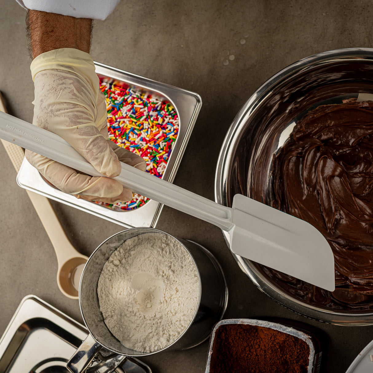 A gloved hand holds a BPA free spatula over bowls of chocolate batter, flour, cocoa powder, and sprinkles on a gray countertop. A wooden spoon and measuring cup complete this inviting baking preparation scene.