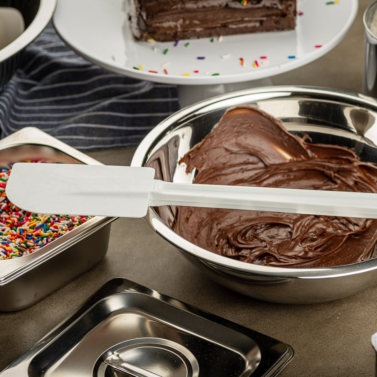 A white, BPA free spatula rests on a metal bowl filled with chocolate cake batter. To the left is a container of colorful sprinkles, and in the background, a plate with partially sliced chocolate cake topped with sprinkles. A striped cloth is partly visible.