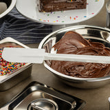 A white, BPA free spatula rests on a metal bowl filled with chocolate cake batter. To the left is a container of colorful sprinkles, and in the background, a plate with partially sliced chocolate cake topped with sprinkles. A striped cloth is partly visible.