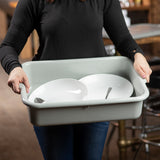 A person in a black long-sleeve shirt holds a large, gray stackable tote box containing two white bowls and a spoon. The person’s face is not visible. The background is blurred, showing indoor furniture.