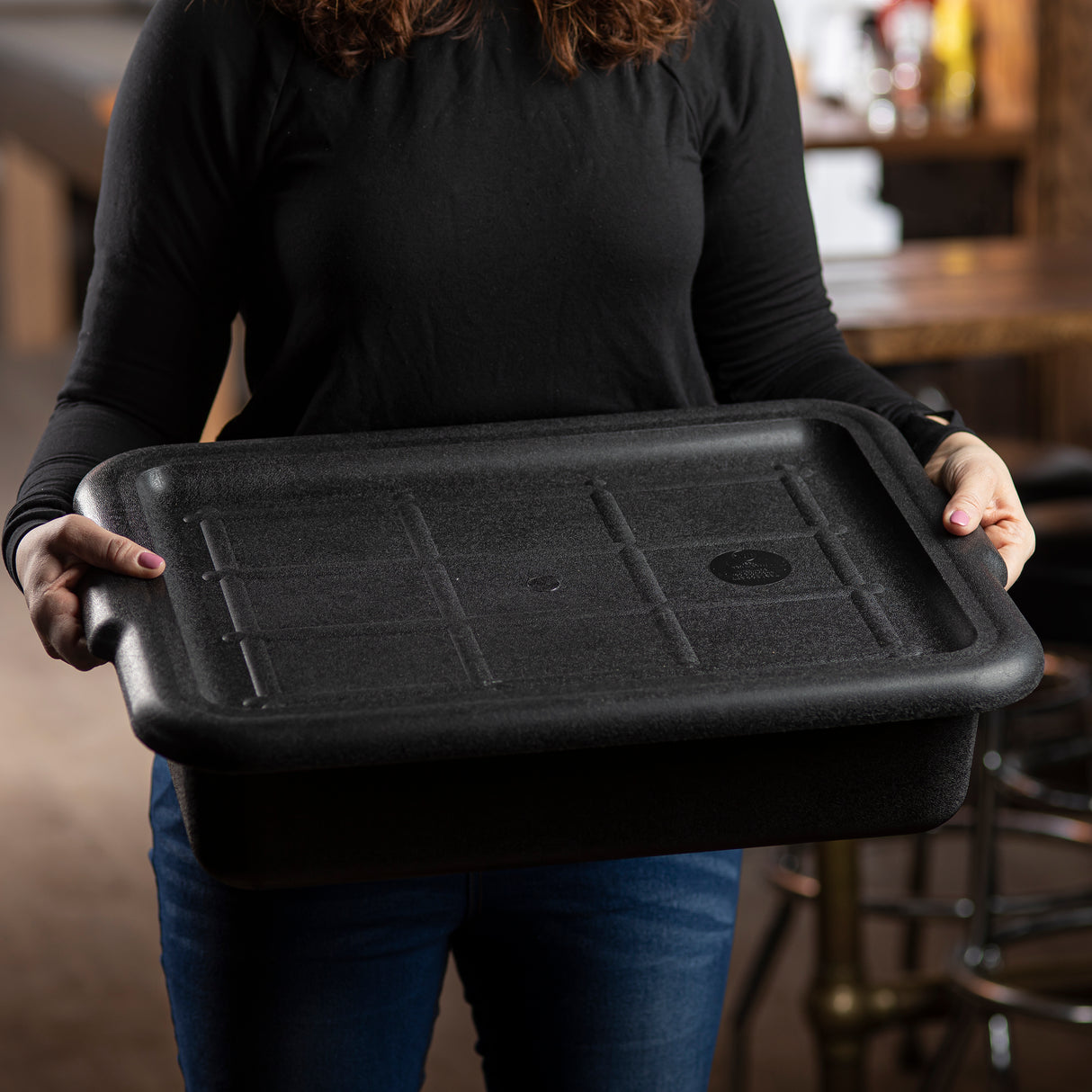 A person in a long-sleeve black shirt and blue jeans holds a large, NSF dishwasher safe food storage container with handles, standing indoors near a wooden table and chairs in a softly lit setting.
