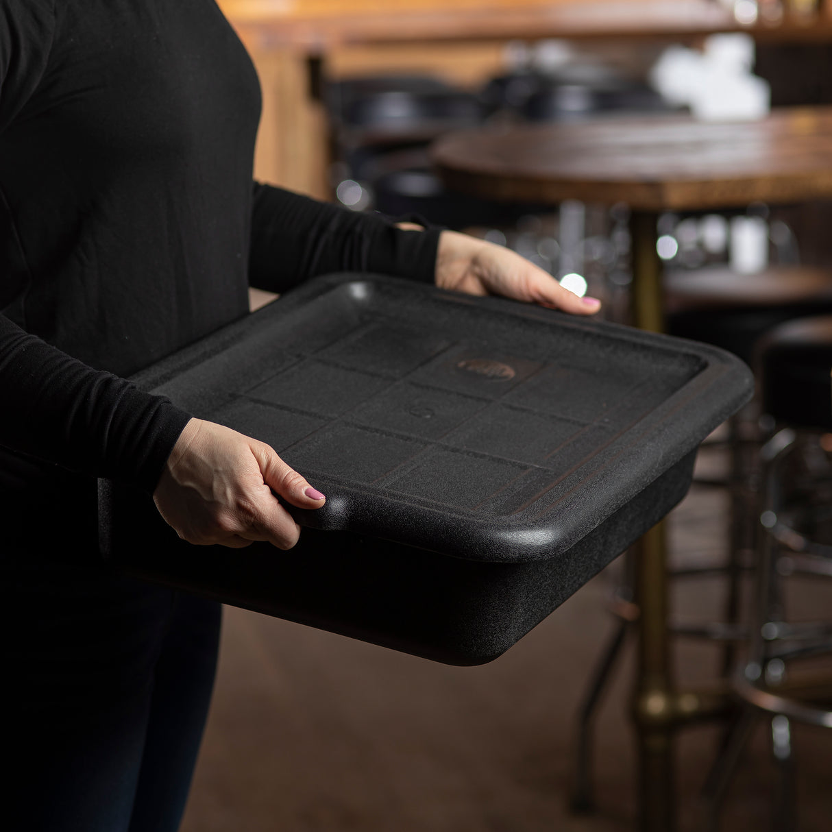 A person in a black long-sleeve shirt holds a large, empty, NSF black rectangular serving tray with both hands. The background shows a wooden table and barstools in a dimly lit dining or restaurant setting.