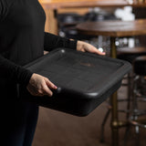 A person in a black long-sleeve shirt holds a large, empty, NSF black rectangular serving tray with both hands. The background shows a wooden table and barstools in a dimly lit dining or restaurant setting.