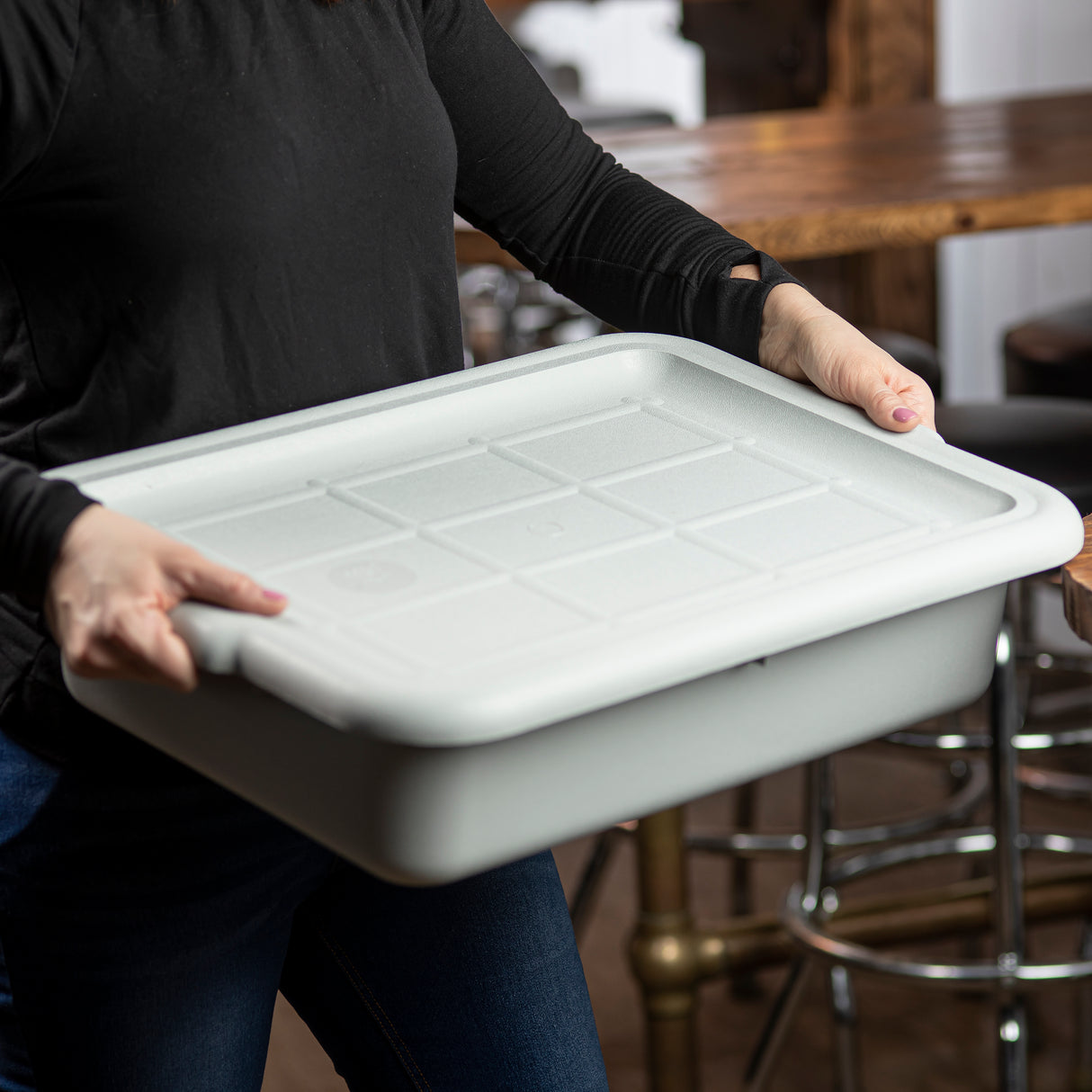 A person in a black long-sleeve shirt and jeans holds a large, rectangular, white storage bin with a fitted Tote Box Cover. The setting is casual, with a wooden table and bar stools. The bin’s durable design is NSF certified and dishwasher safe.