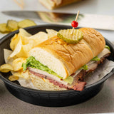 A sub sandwich with lettuce, sliced roast beef, cheese, and turkey on a hoagie roll, topped with pickles and a toothpick, is served in a heat resistant platter basket with potato chips. Sliced pickles and a knife are blurred in the background.