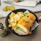 A sandwich with lettuce, cheese, and deli meat on a hoagie roll, topped with a pickle slice and toothpick, sits in a dishwasher safe platter basket lined with parchment paper next to potato chips. A pepper shaker, pickle slices, and knife are in the background.