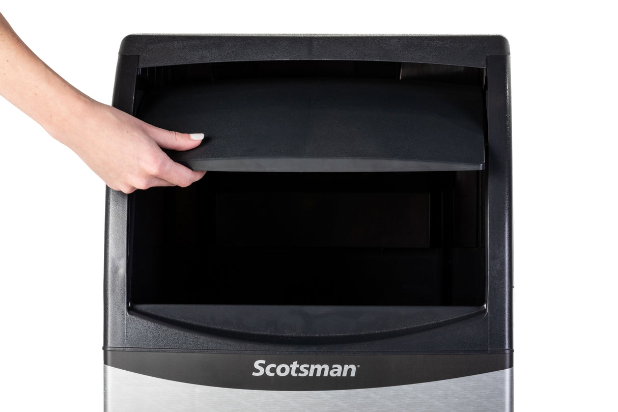 A hand with neatly manicured nails lifts the black door of a Scotsman Ice Maker. The stainless steel undercounter unit displays the Scotsman name, and the open top compartment reveals the interior of this cube style ice maker.