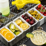 A stainless steel condiment holder with 6 compartments displays orange slices, lime wedges, lemon slices, cherries, green olives, and strawberries. Nearby are a yellow citrus squeezer, a bottle, metal cups, and a creamy drink garnished with cucumber.