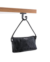 A black designer handbag with a subtle monogram pattern and a short strap hangs from a TableCraft bag hook attached to the underside of a wooden shelf, set against a plain white background.