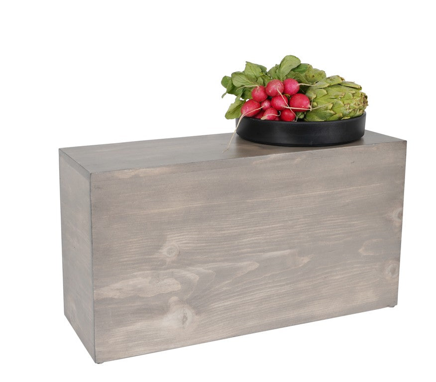 A rectangular Aspen Riser made of light gray-washed pine wood stands upright. On top, a round black bowl holds fresh produce, including artichokes and radishes with leafy greens. The background is plain white, highlighting the Cal-Mil design.