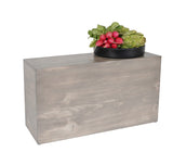 A rectangular Aspen Riser made of light gray-washed pine wood stands upright. On top, a round black bowl holds fresh produce, including artichokes and radishes with leafy greens. The background is plain white, highlighting the Cal-Mil design.