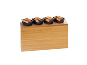 A rectangular Cal-Mil bamboo riser with four small loaves of bread in dark paper liners neatly arranged in a row on top, all against a plain white background.
