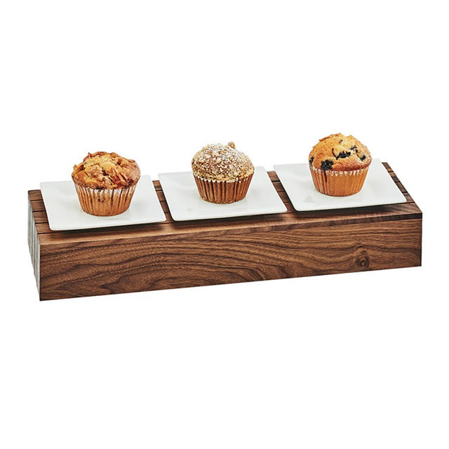 Three muffins rest on individual square white plates, placed on a rectangular walnut display tray. Each muffin features a unique topping or flavor, with one dusted in powdered sugar. The background is plain white.
