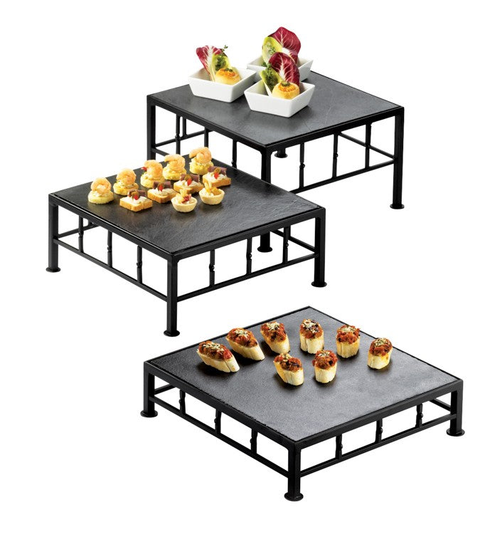 Three black square iron frame risers of varying heights display appetizers: small cups with leafy greens on the tallest, bite-sized items topped with herbs in the middle, and oval-shaped pieces with garnishes on the lowest, all against a white background.