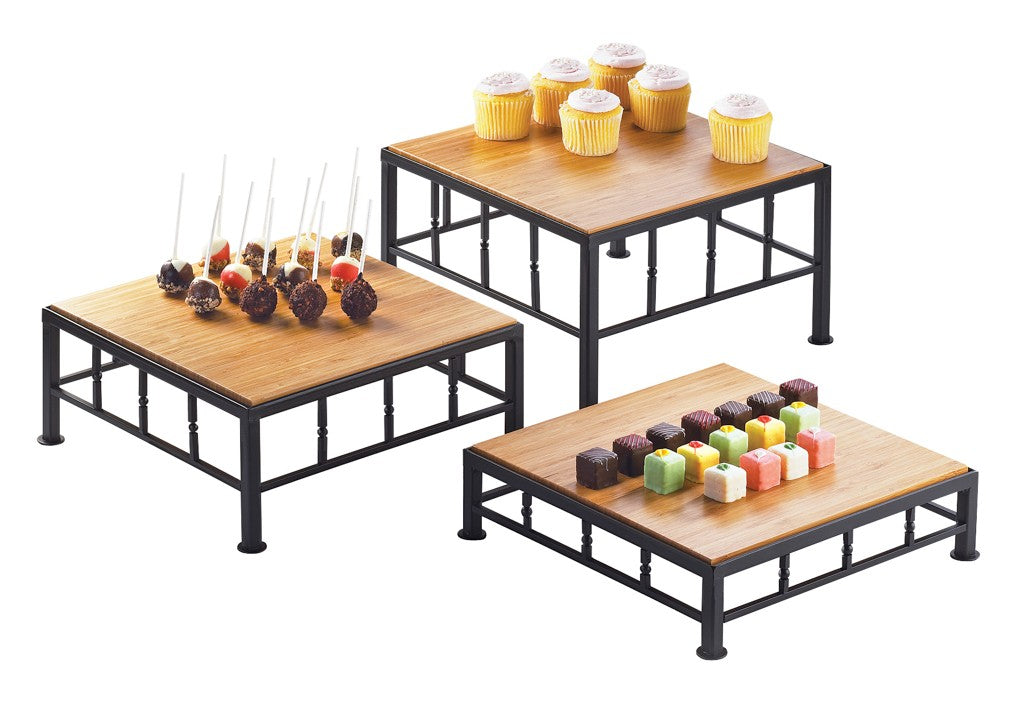 Three black metal and wood display risers, each a different height, hold desserts: six yellow cupcakes with white frosting, assorted cake pops with sticks, and colorful square petit fours. Each riser features a bamboo top and sturdy iron frame on a white background.