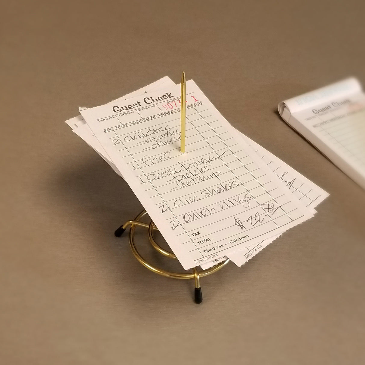 A stack of handwritten restaurant guest checks is skewered on a brass plated check spindle from TableCraft Products, resting on a beige surface. The visible check lists food items and prices, with another pad partially visible in the background.