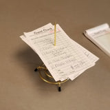 A stack of handwritten restaurant guest checks is skewered on a brass plated check spindle from TableCraft Products, resting on a beige surface. The visible check lists food items and prices, with another pad partially visible in the background.