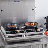 Two frying pans sit on an induction cooktop with digital controls; the left pan holds steak and garlic, while the right contains a colorful vegetable medley. Carrots and stacked black pots rest nearby in a spotless, stainless steel kitchen setting.