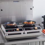 Two induction cooktops on a stainless steel counter, each with a non-stick fry pan; the left pan has steaks and garlic, the right sizzles with a colorful vegetable medley. Digital displays glow as carrots and black pots rest nearby.