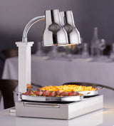 A carving station with an adjustable neck double heat lamp shines over sliced roast meat and cheesy macaroni. The stainless steel setup sits on a white tablecloth, with blurred tables and chairs hinting at a restaurant or banquet setting.