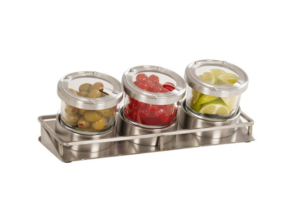 Three glass jars with stainless steel lids sit in a cold condiment display tray. The jars contain green olives, red cherries, and lemon or lime wedges, arranged side by side and slightly tilted forward for easy access.