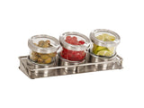 Three glass jars with stainless steel lids sit in a cold condiment display tray. The jars contain green olives, red cherries, and lemon or lime wedges, arranged side by side and slightly tilted forward for easy access.
