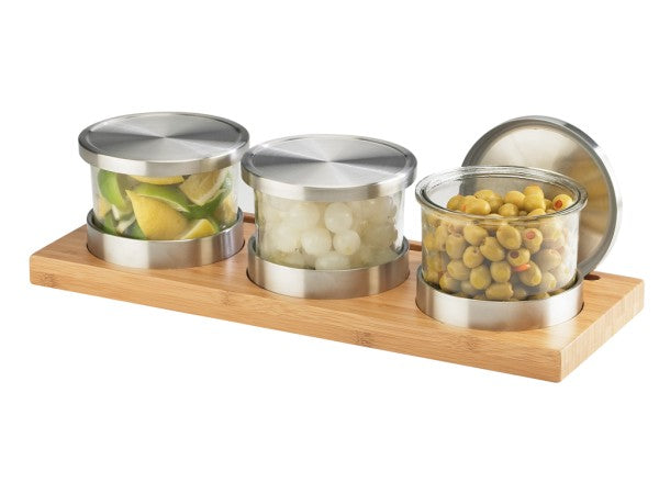 Three glass jars with silver lids sit on a rectangular bamboo base, perfect for a Cold Condiment Display. The jars hold lime wedges, pearl onions, and green olives with pimentos. One lid is leaning upright at the back right of the tray.