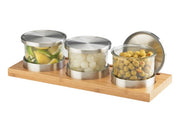 Three glass jars with silver lids sit on a rectangular bamboo base, perfect for a Cold Condiment Display. The jars hold lime wedges, pearl onions, and green olives with pimentos. One lid is leaning upright at the back right of the tray.