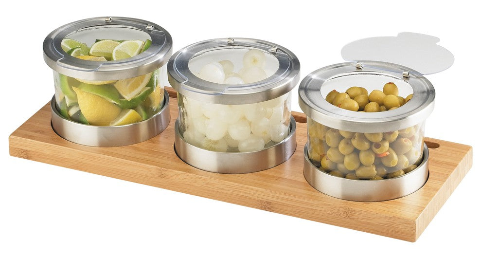 Three round glass jars with metal lids rest on a rectangular bamboo base. The cold condiment display holds lime wedges, cocktail onions, and green olives. One lid is propped open, showing the olives inside—perfect for serving garnishes.
