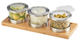Three round glass jars with metal lids rest on a rectangular bamboo base. The cold condiment display holds lime wedges, cocktail onions, and green olives. One lid is propped open, showing the olives inside—perfect for serving garnishes.