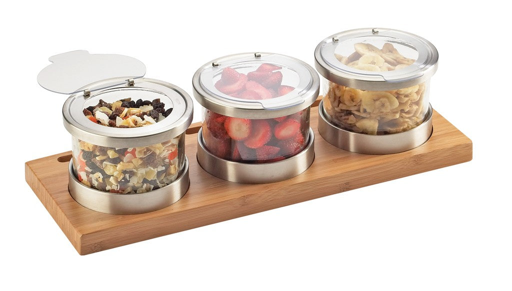 Three glass jars with stainless steel lids sit on a bamboo base. The jars contain diced dried fruit mix, sliced strawberries, and banana chips, with lids partially open for a vibrant cold condiment display.