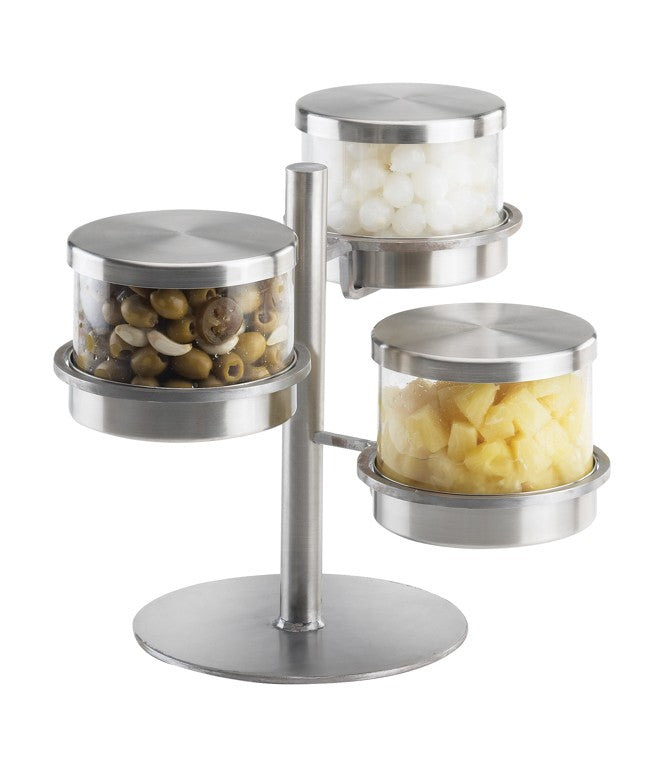 A tiered, rotating stainless steel Mixology Condiment Display with three clear, round glass jars. Each jar has a metal lid and holds green olives, pearl onions, and cubed pineapple. The stand sits on a circular base.