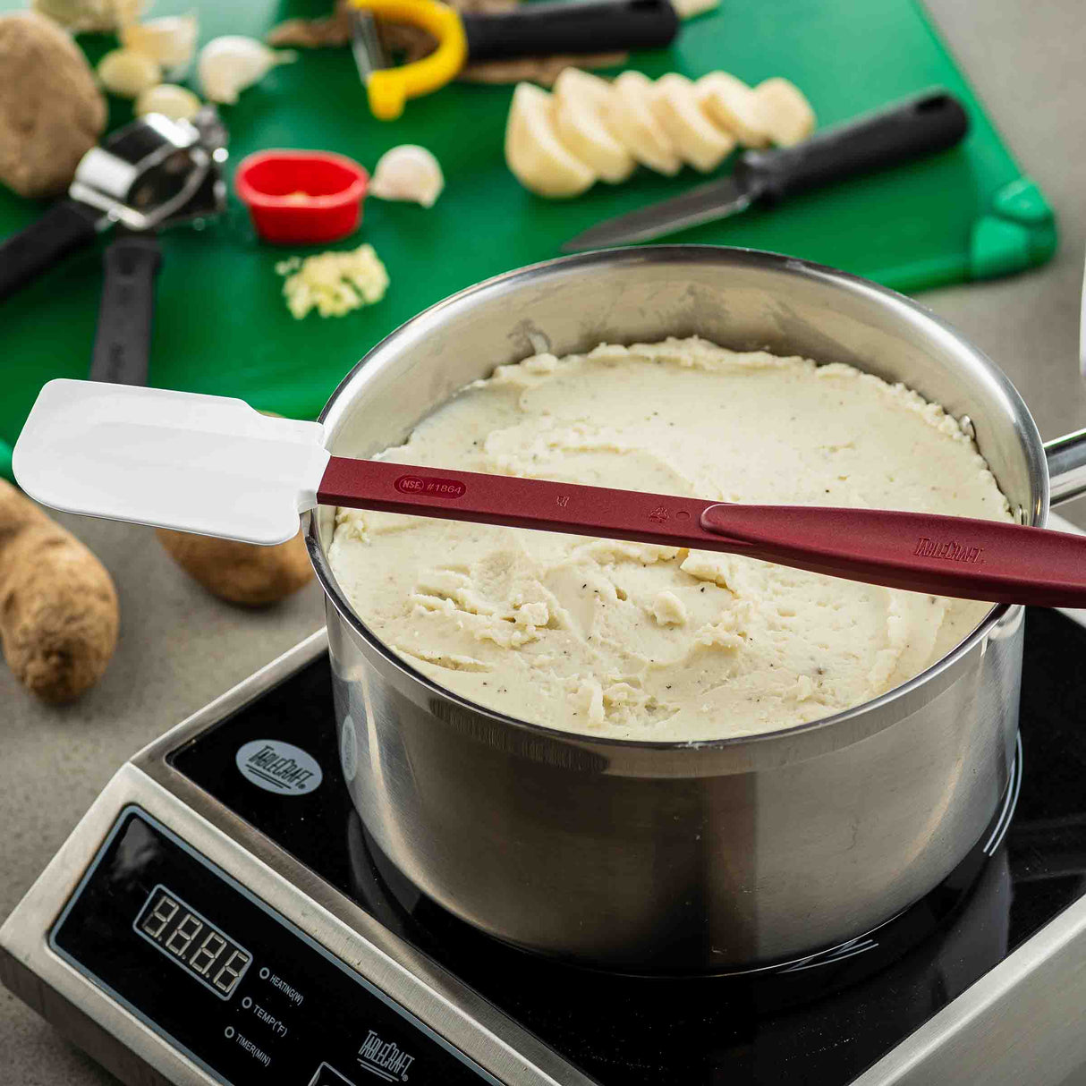 A pot of mashed potatoes sits on an electric stovetop. A red and white high heat spatula rests across the pot. In the background, a green cutting board holds sliced garlic, a yellow peeler, a small red measuring cup, and unpeeled potatoes.