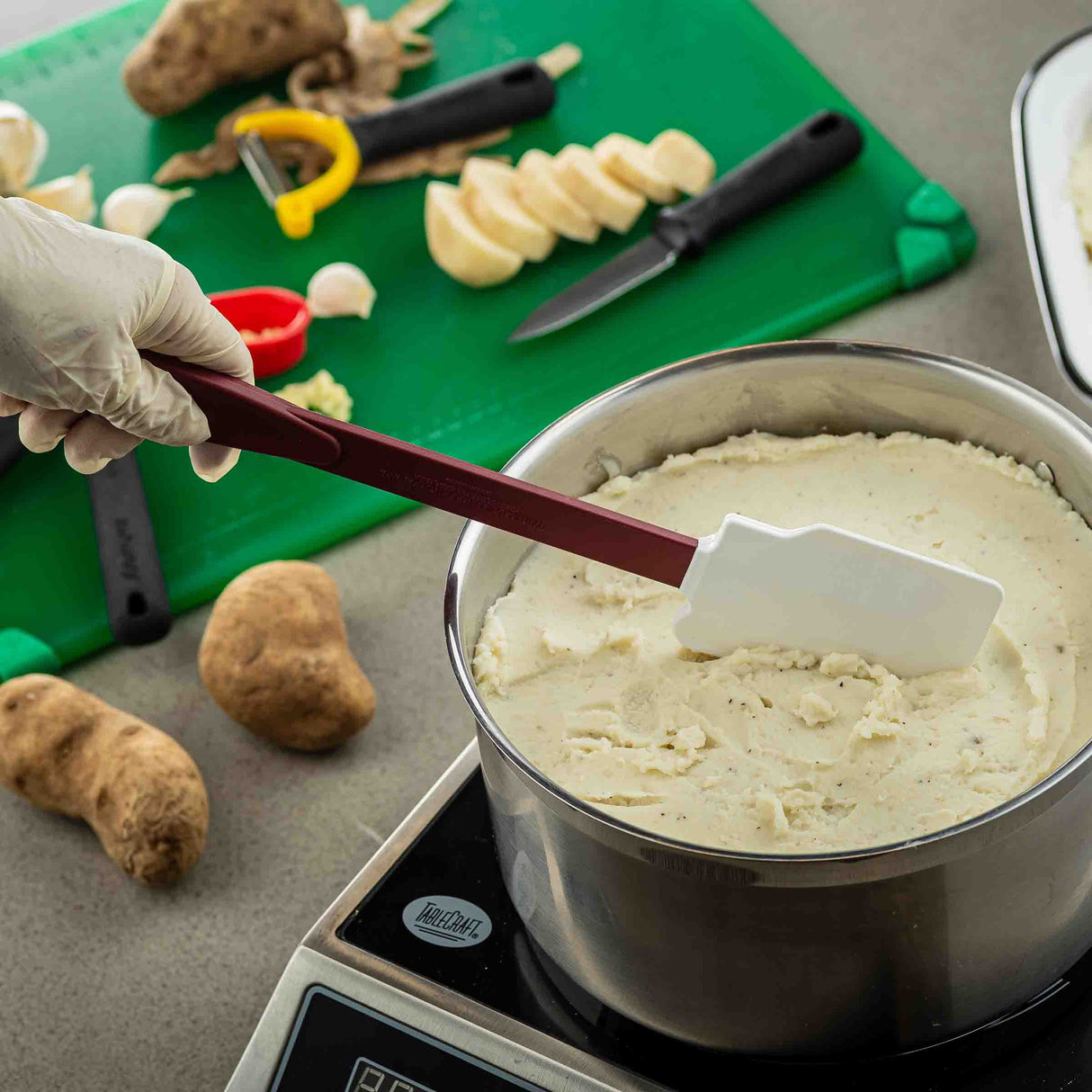 A hand wearing a glove holds a BPA free silicone spatula, stirring creamy mashed potatoes in a pot on a stovetop. In the background, whole potatoes, peeled slices, a vegetable peeler, knives, and a green cutting board are visible.