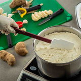 A hand wearing a glove holds a BPA free silicone spatula, stirring creamy mashed potatoes in a pot on a stovetop. In the background, whole potatoes, peeled slices, a vegetable peeler, knives, and a green cutting board are visible.