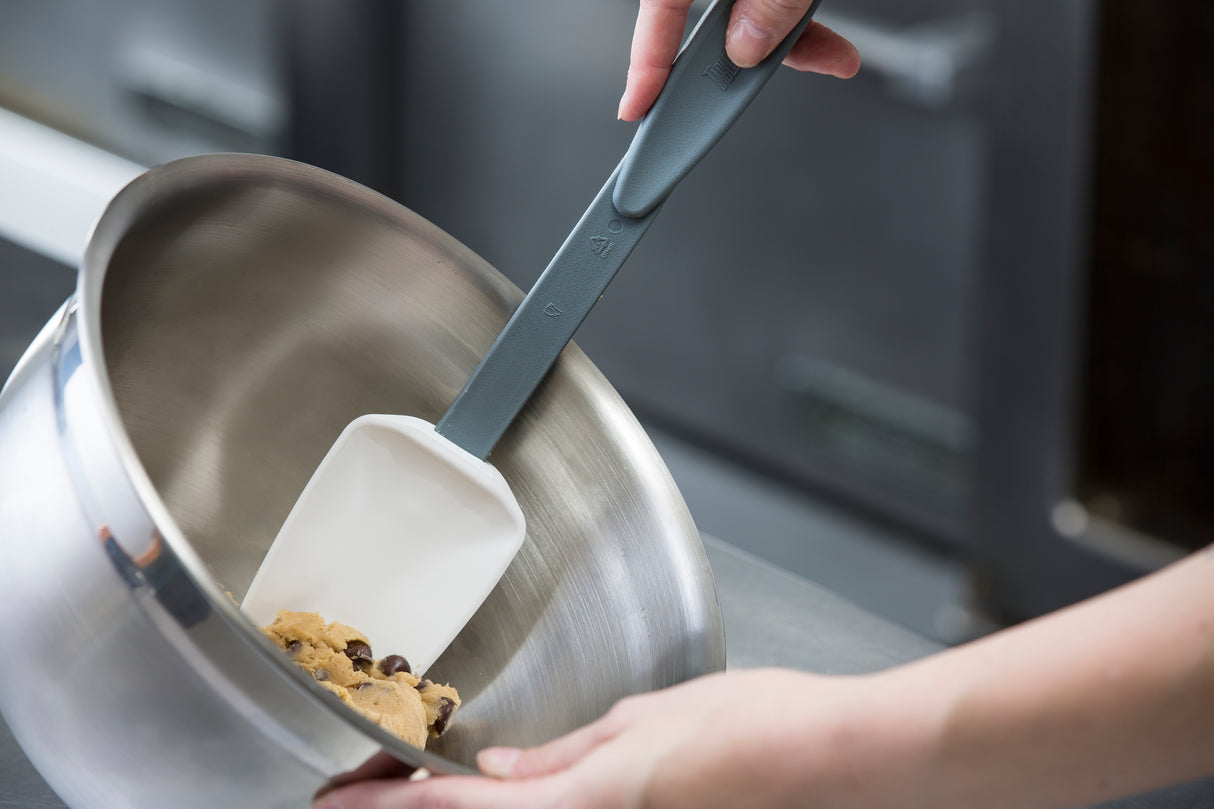 A close-up of hands using a BPA free, high heat resistant silicone spoon to mix chocolate chip cookie dough in a stainless steel mixing bowl, with a blurred kitchen background.