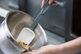 A close-up of hands using a BPA free, high heat resistant silicone spoon to mix chocolate chip cookie dough in a stainless steel mixing bowl, with a blurred kitchen background.
