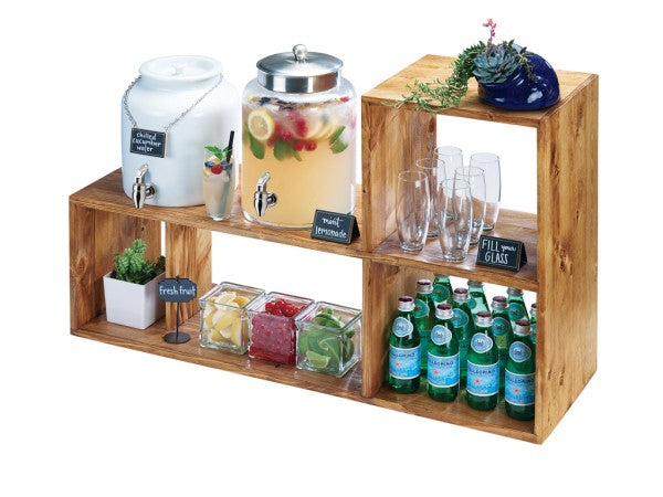 A rustic pine drink station with a removable shelf holds a white water dispenser, lemonade dispenser, small plant, drinks, fruit cubes, and blackboard labels. The setup is organized in open cube shelves against a white background.