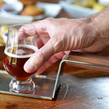 A close-up of a hand reaching for a small glass of amber beer, placed on a stainless steel tray with a wooden handle. In the blurred background, plates of food are visible on a wooden table, suggesting a casual dining setting.