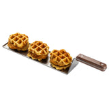 Three pieces of fried chicken sandwiched between round golden waffles are arranged in a row on the stainless steel Slider Flight Spatula with a brown wooden handle, set against a plain white background.