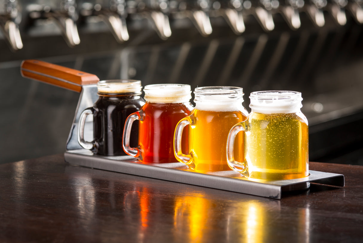 A stainless steel plate holds four mason jar mugs of beer in varying colors, arranged in a row on a wooden table. Behind the tray, several beer taps are visible out of focus, suggesting a lively bar setting. Perfect for a TableCraft Products drink flight.