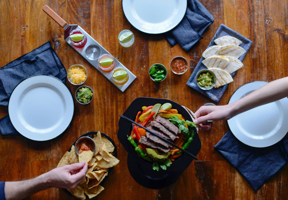 Overhead view of a wooden table set for a meal. Plates, napkins, and drinks—including a Drink flight spatula—surround fajita steak with grilled peppers, tortillas, cheese, salsa, guacamole, jalapeños, chips, and two people reaching for food.