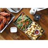A tabletop view of stainless steel trays with food: buffalo chicken wings, a fresh spinach salad topped with shredded carrots, and crispy tater tots with ketchup. A dish of ranch dressing and salt and pepper shakers are also visible on the wooden table.