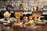 A wooden table set with burgers from a Burger Box topped with lettuce, tomato, and cheese, sliders, fries, loaded tots, grilled vegetables, and two pints of beer—all served in dishwasher safe stainless steel trays; a blurred bar is in the background.