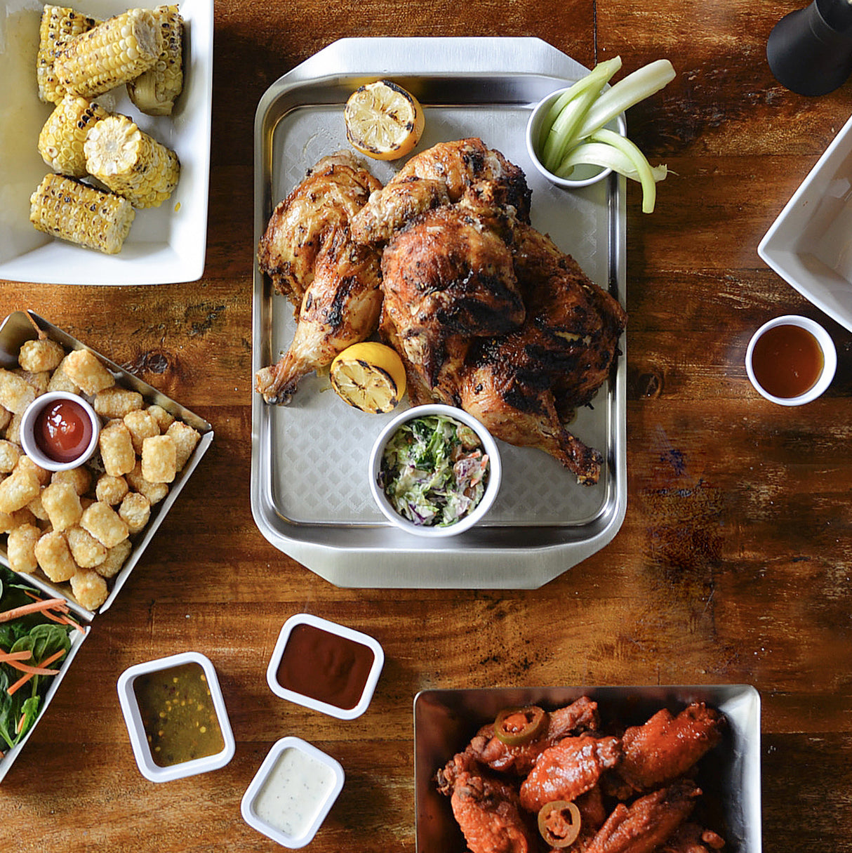 A serving tray of roasted chicken with lemon halves and coleslaw is at the center. Surrounding it are corn on the cob, tater tots with ketchup, celery sticks with dip, sauced chicken wings, and salad from the Better Burger Collection on a wooden table.