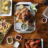 A serving tray of roasted chicken with lemon halves and coleslaw is at the center. Surrounding it are corn on the cob, tater tots with ketchup, celery sticks with dip, sauced chicken wings, and salad from the Better Burger Collection on a wooden table.