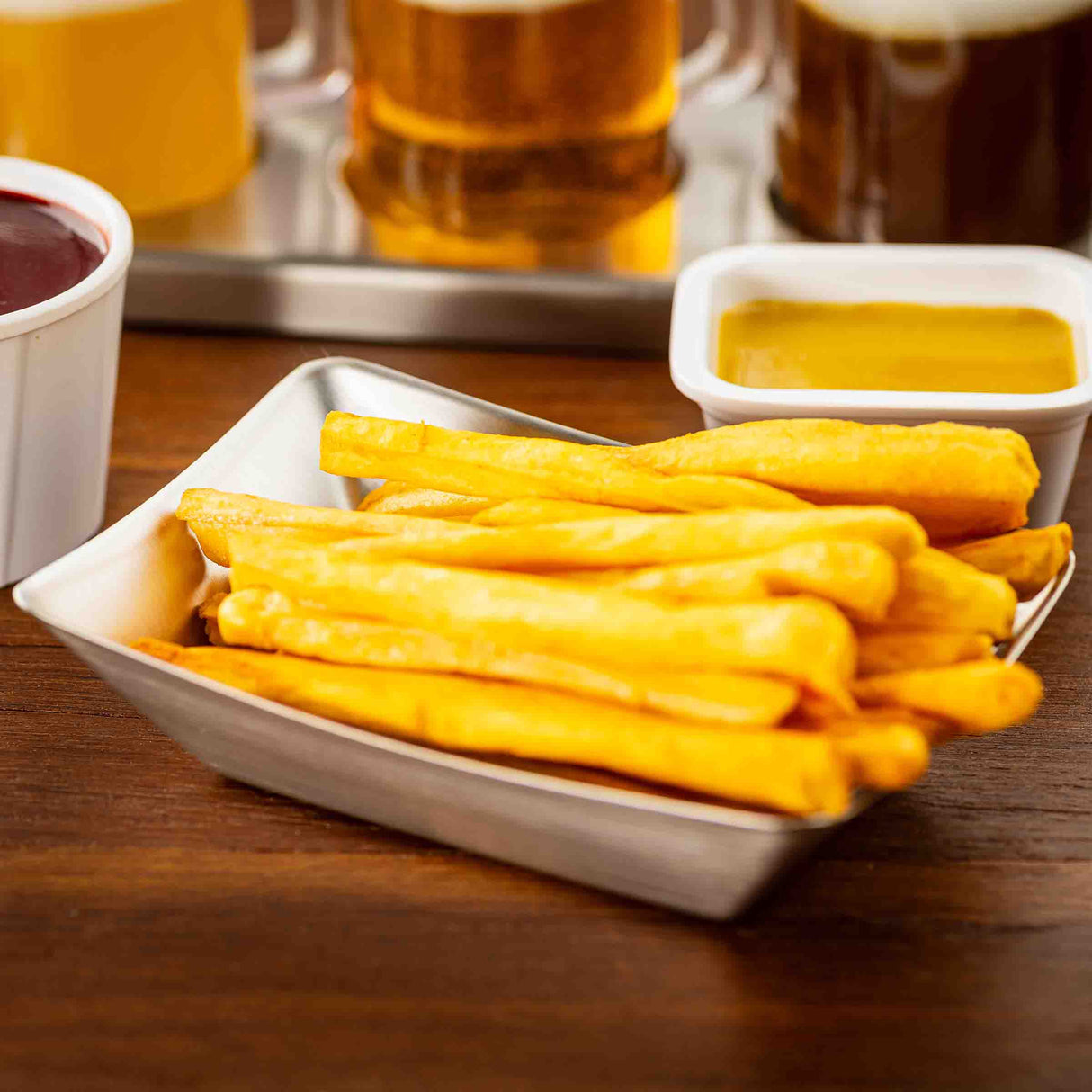 A stainless steel fry tray filled with golden French fries sits on a wooden table, accompanied by ketchup and mustard. In the background, three glasses of beer are partially visible, suggesting a casual dining or bar setting.
