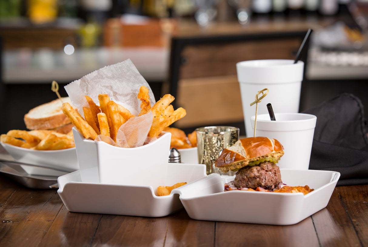 A close-up of a meal from the Better Burger Collection featuring golden fries in a white bowl, a small burger with lettuce on a brioche bun, and a white cup with a straw, all on a wooden table. The background is blurred, showing more food and drinks.